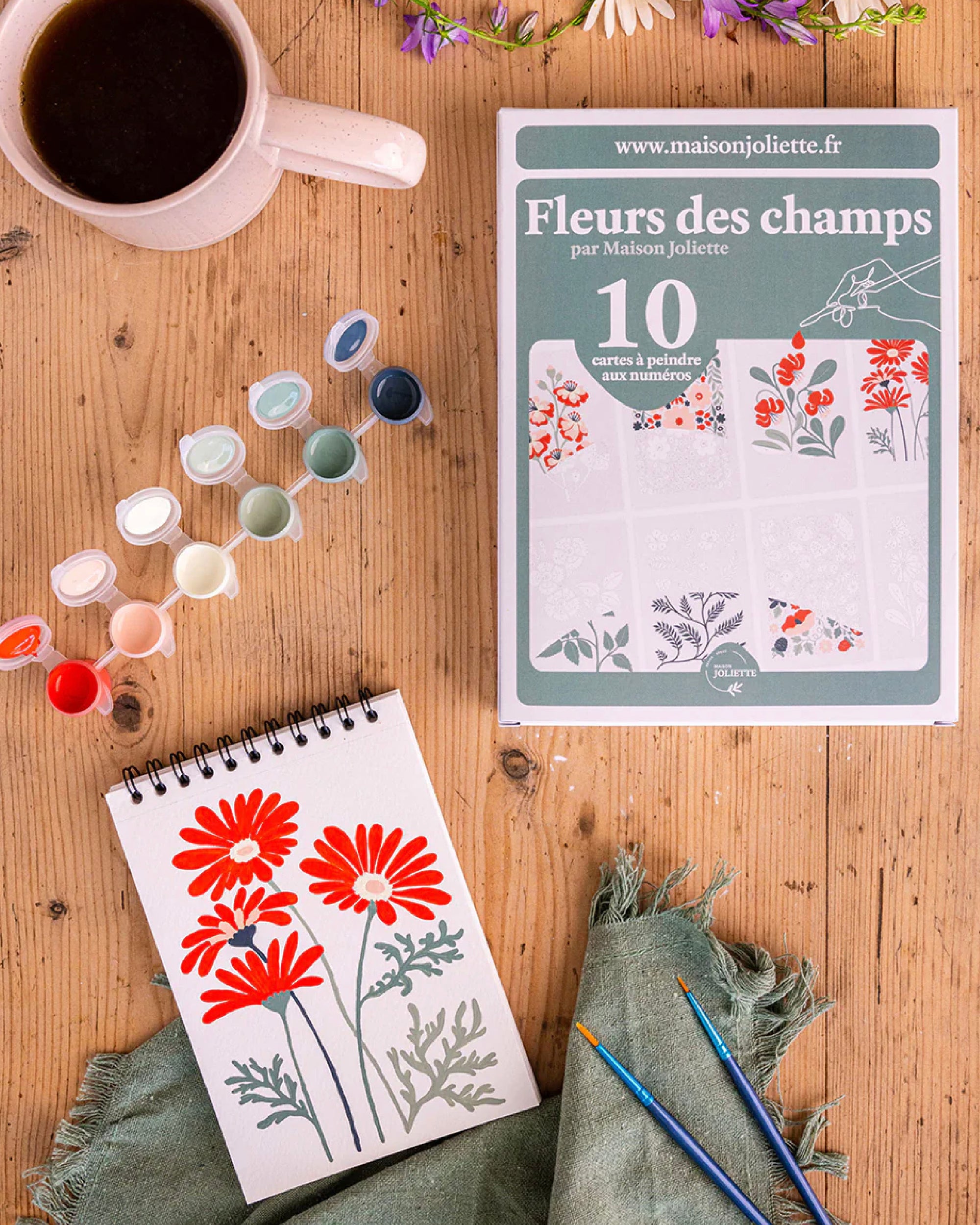 French paint by numbers craft kit box on a wooden table with a paint set, paintbrushes, sketchbook with flowers and a coffee mug