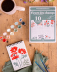French paint by numbers craft kit box on a wooden table with a paint set, paintbrushes, sketchbook with flowers and a coffee mug