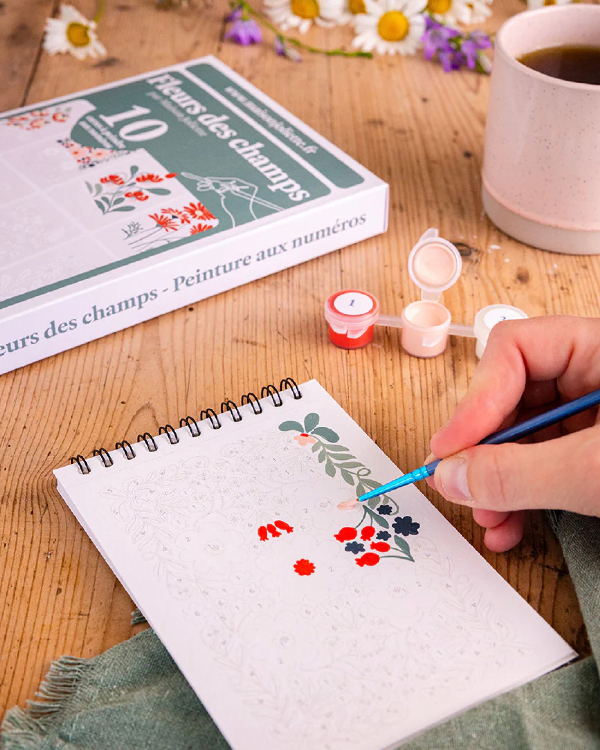 Close up of a person painting on a small Fleurs des champs Frenchi painging kit sketchbook with a cup of coffee and art supplies in the background
