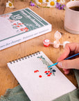 Close up of a person painting on a small Fleurs des champs Frenchi painging kit sketchbook with a cup of coffee and art supplies in the background
