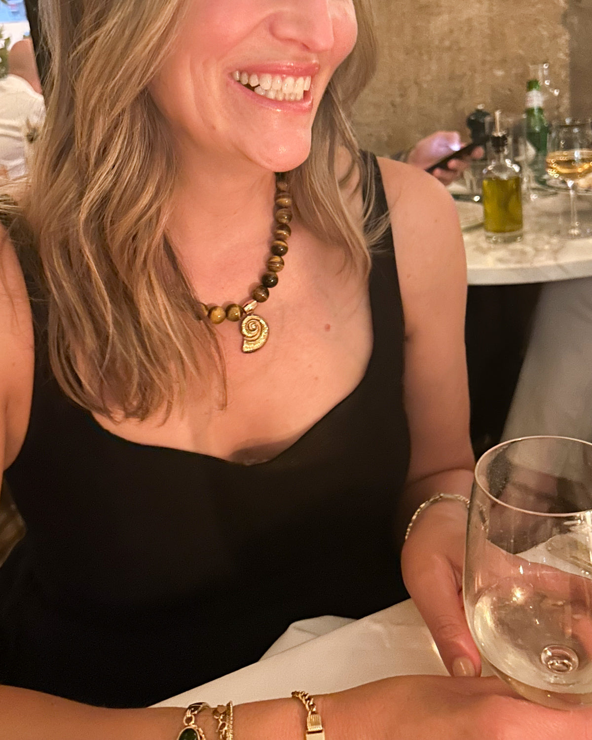 A woman in a black sleeveless top smiling while wearing a gold pendant necklace and bracelets, holding a clear wine glass in a restaurant setting.