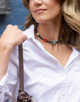 A close-up lifestyle shot of a woman wearing a colorful beaded choker necklace with a white button-down shirt. The necklace features dark blue beads with circular accents in red, light blue, yellow, and white.