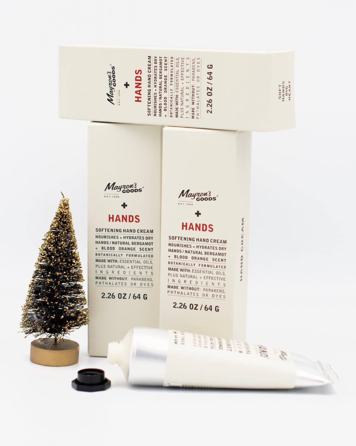Opened tube of Mayron's Goods Hand Cream in front of 3 boxes next to a mini bristle Christmas tree. 