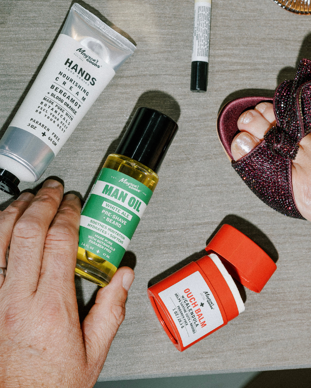 Man body care products including a tube of hand cream, bottle of man oil, and stick of ouch balm ointment on a table.