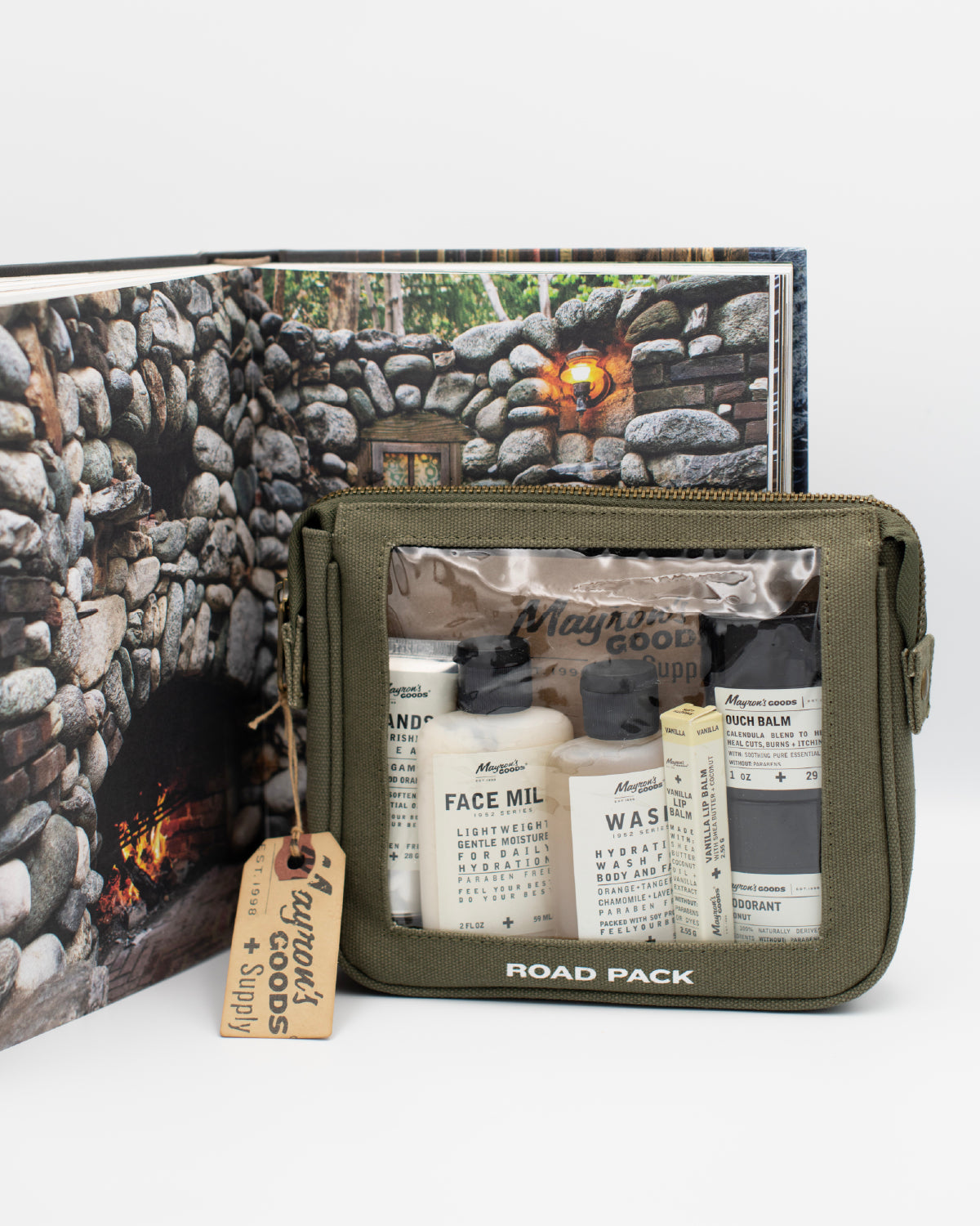 Army green travel pack with Mayron's Goods products in front of an opened book with a stone fireplace image.