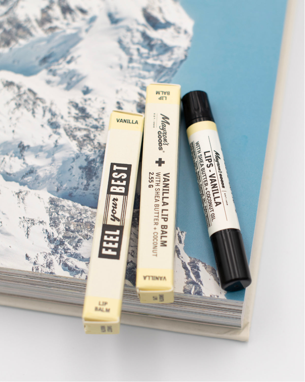 Close up of a tube of Mayron&#39;s Goods Vanilla Lip Balm next to two packages on an opened book fearuting a snowy mountain.