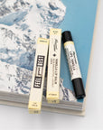Close up of a tube of Mayron's Goods Vanilla Lip Balm next to two packages on an opened book fearuting a snowy mountain.