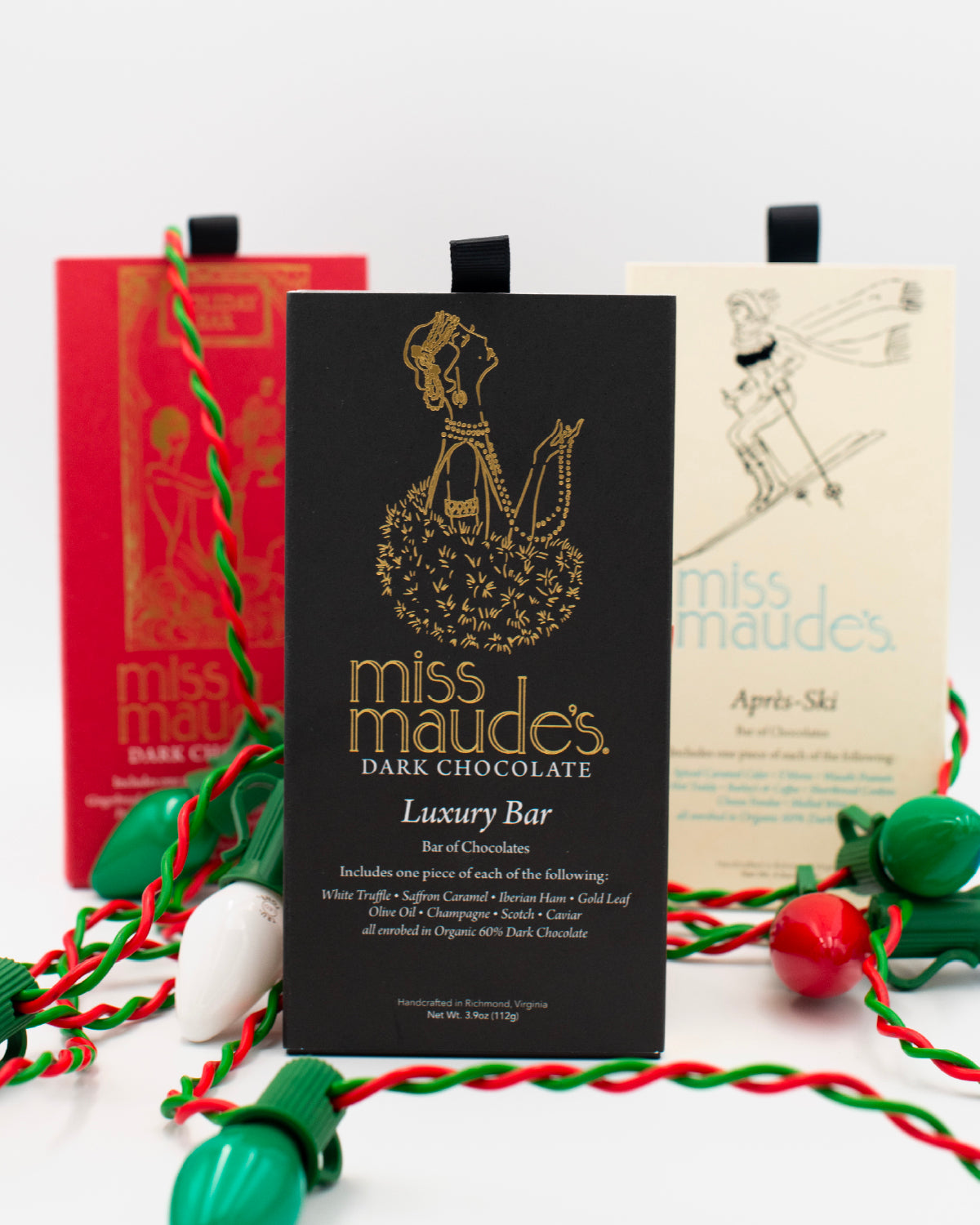 Three, Miss Maude's dark chocolate bars: Luxury Bar, Holiday Bar, Aprés Ski bar standing upright with festive green and red holiday lights strewn around them.