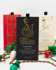 Three, Miss Maude's dark chocolate bars: Luxury Bar, Holiday Bar, Aprés Ski bar standing upright with festive green and red holiday lights strewn around them.