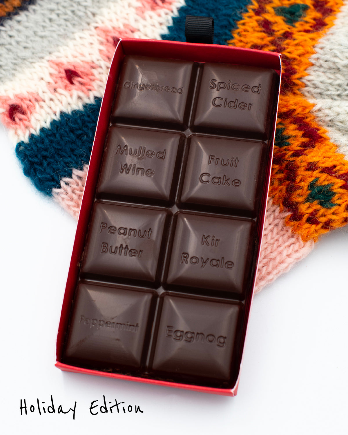 Close-up of an opened Miss Maude’s Holiday Edition dark chocolate bar with eight flavored squares displayed on a patterned sweater background.