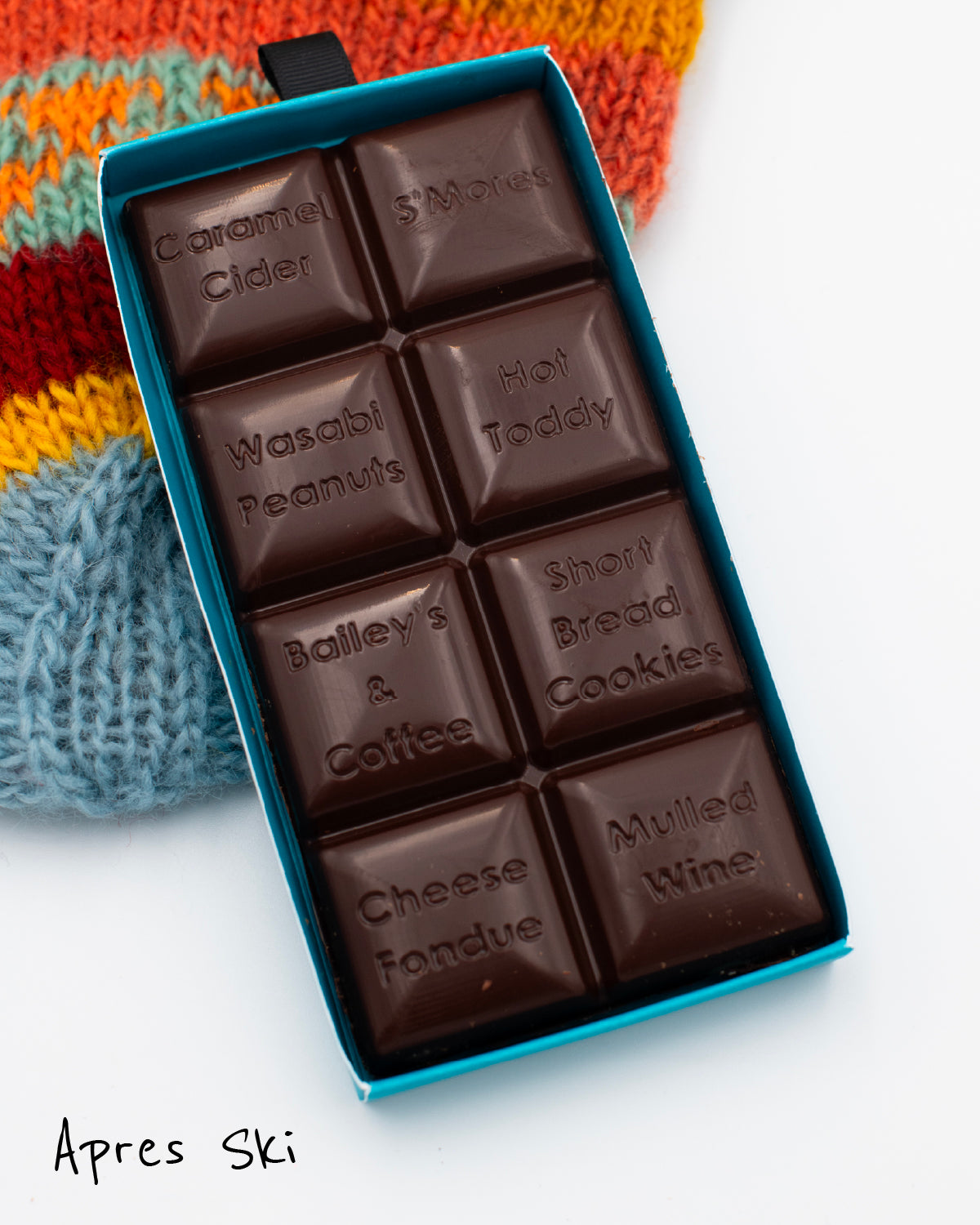 Close-up of an opened Miss Maude’s Aprés Ski dark chocolate bar with eight flavored squares displayed on a colorful knit hat 