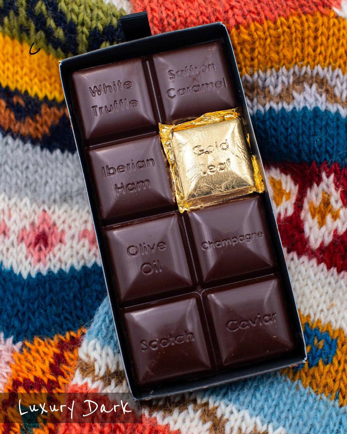 Close-up of an opened Miss Maude’s Luxury Bar dark chocolate bar with eight flavored squares, including one wrapped in gold foil, displayed on a patterned sweater background.