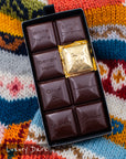 Close-up of an opened Miss Maude’s Luxury Bar dark chocolate bar with eight flavored squares, including one wrapped in gold foil, displayed on a patterned sweater background.