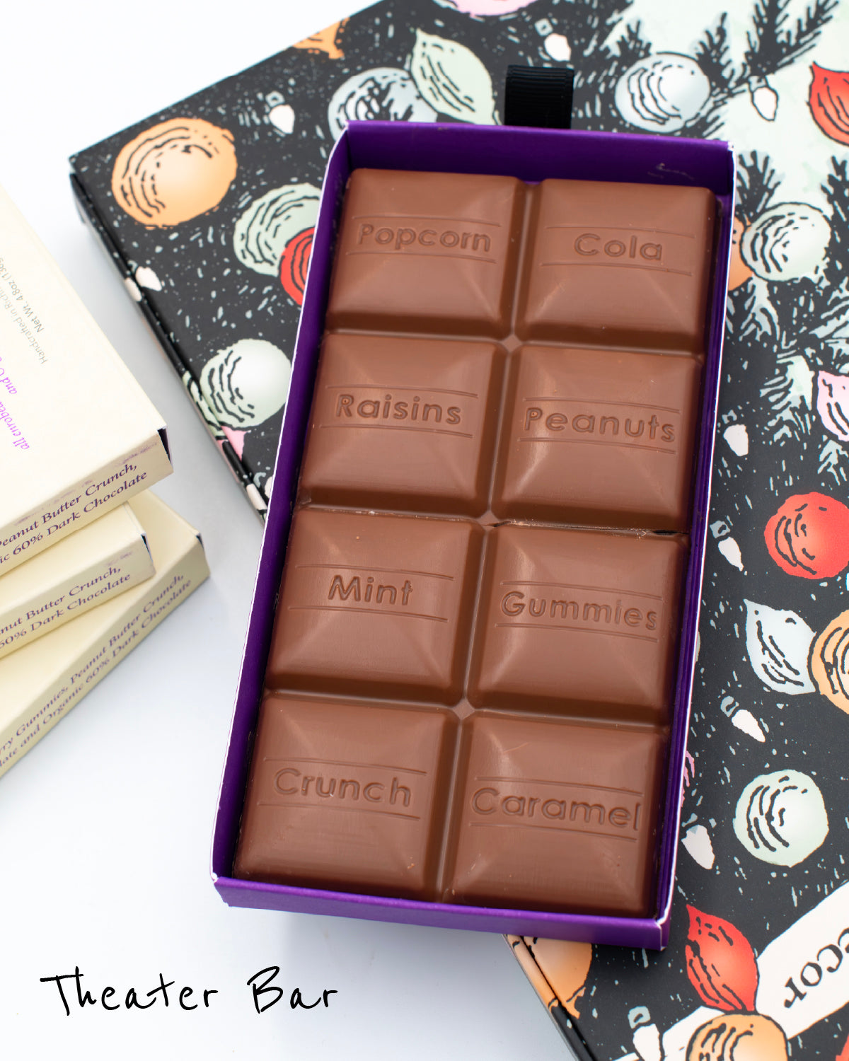 Close up of opened box of Miss Maude&#39;s Theater Bar featuring 8 milk chocolate squares in different flavors on a holiday illustration background