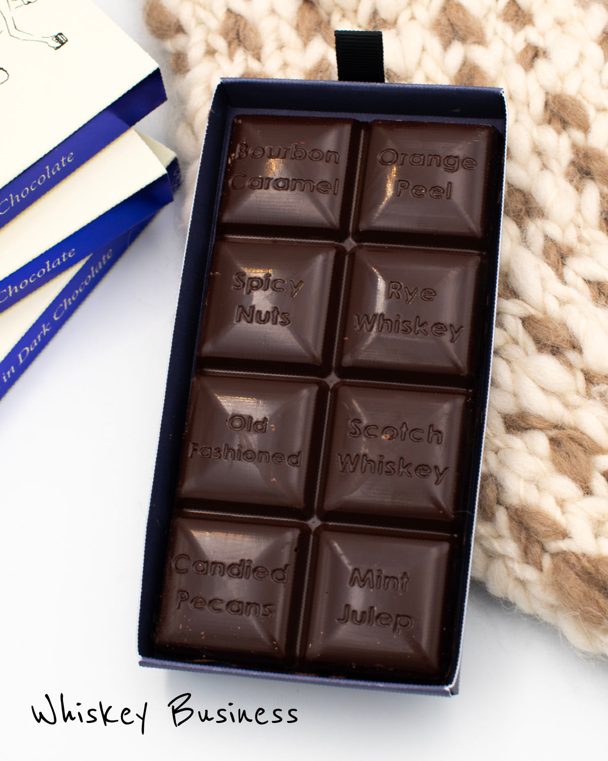 Close up of open box of Miss Maude&#39;s Whiskey Business dark chocolate bar, featuring 8 squares with difffent flavors on a white and beige knit background 