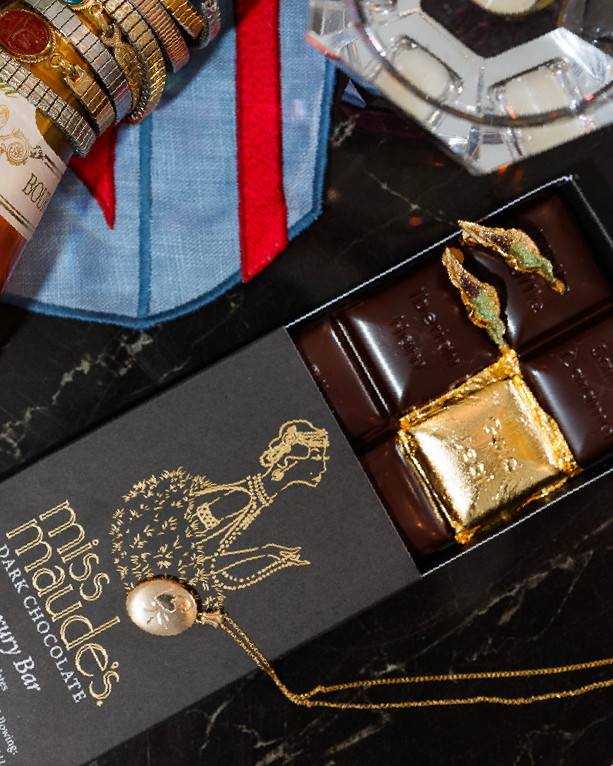 Partially opened box of Miss Maude'Luxury Bar: dark chocolate gold foil wrapping on one square with a gold necklace laying on the box