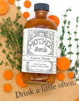 Bottle of 'Mother Shrub Carrot Thyme' with herbs and carrots on a wooden surface.