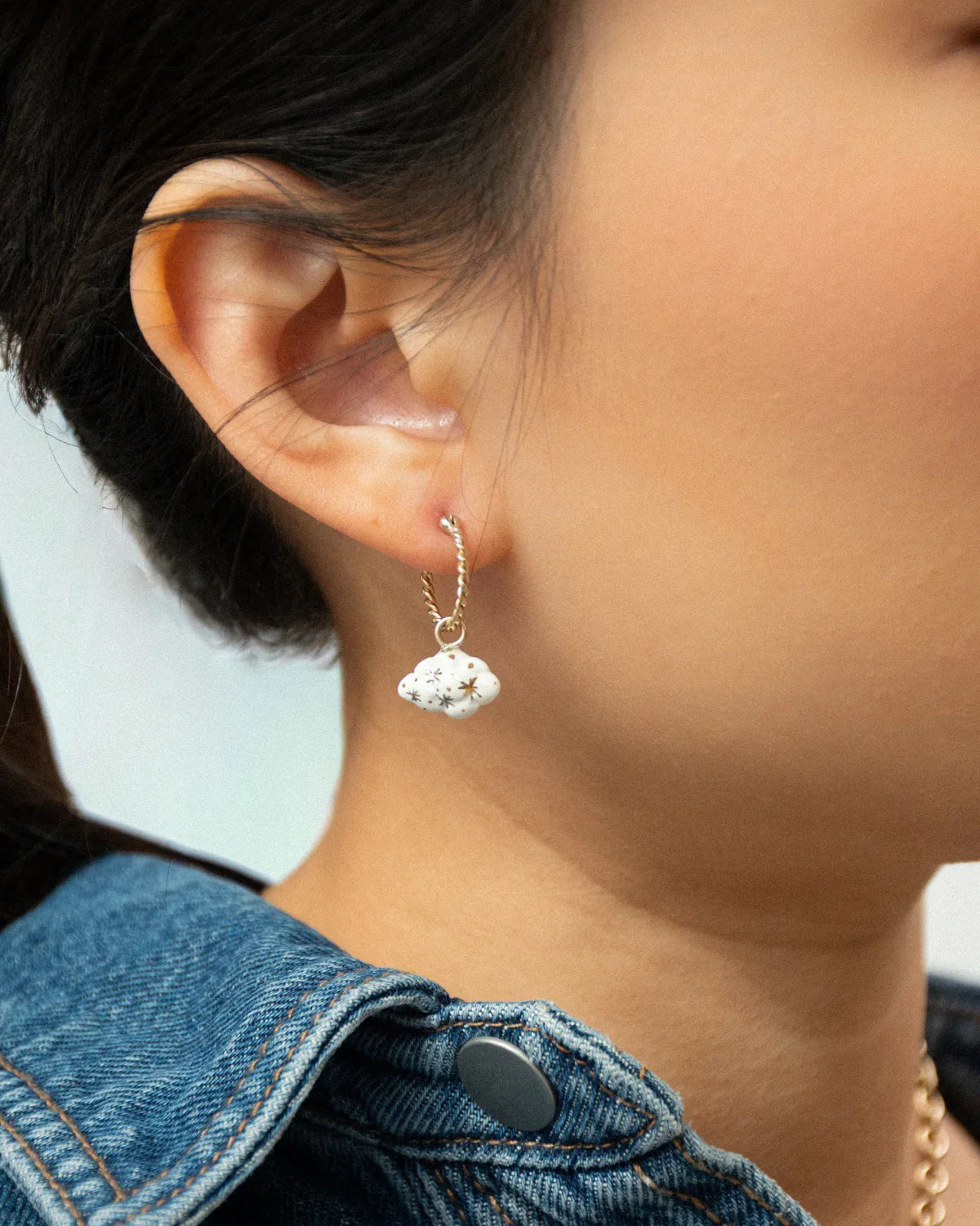 Close-up of an ear wearing a Daydreamer gold hoop with a white cloud charm, with a denim jacket