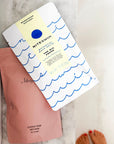 Niteswim The Big Pause bath salts pouch with a wave print, lying on a pink Merigold pouch