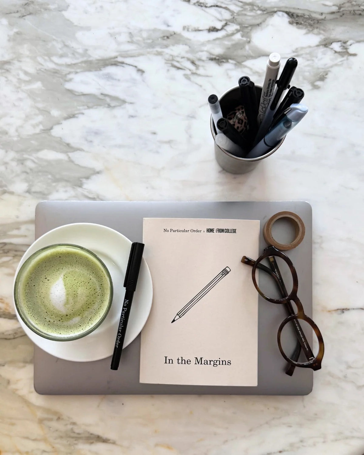 In the Margins journal on a laptop beside a matcha latte, glasses and pens on marble