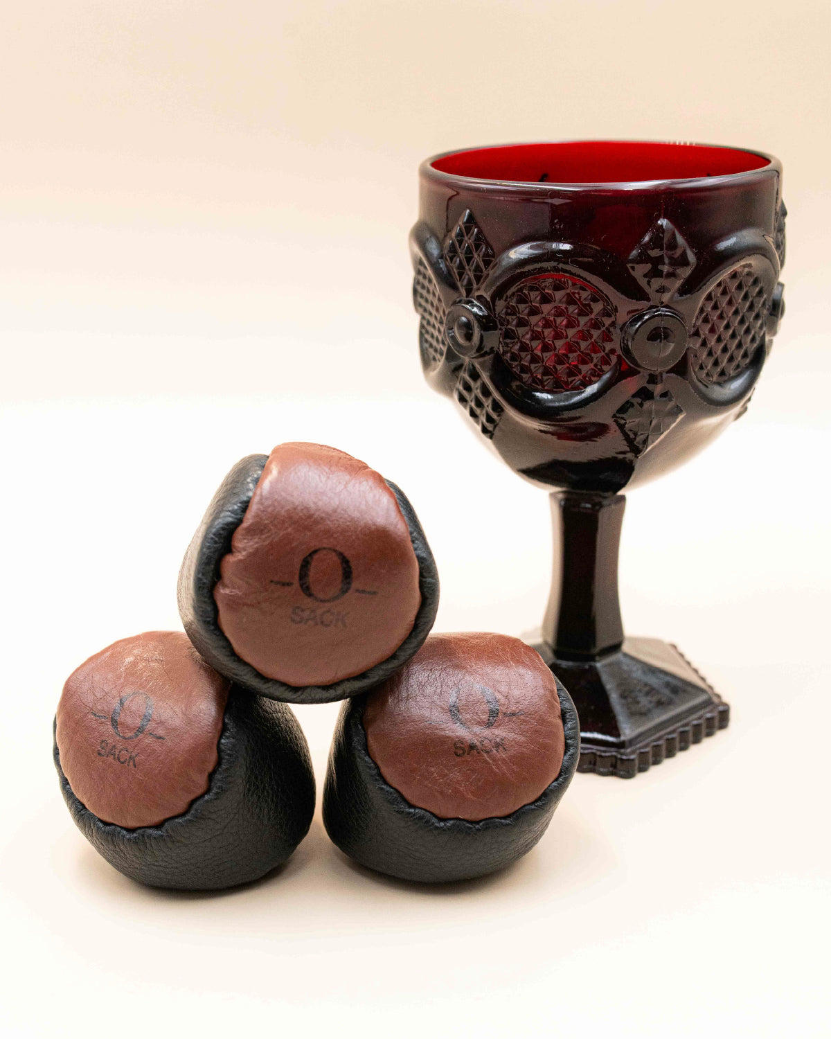 Three brown leather O-Sack hacky sacks stacked in front of a red glass goblet