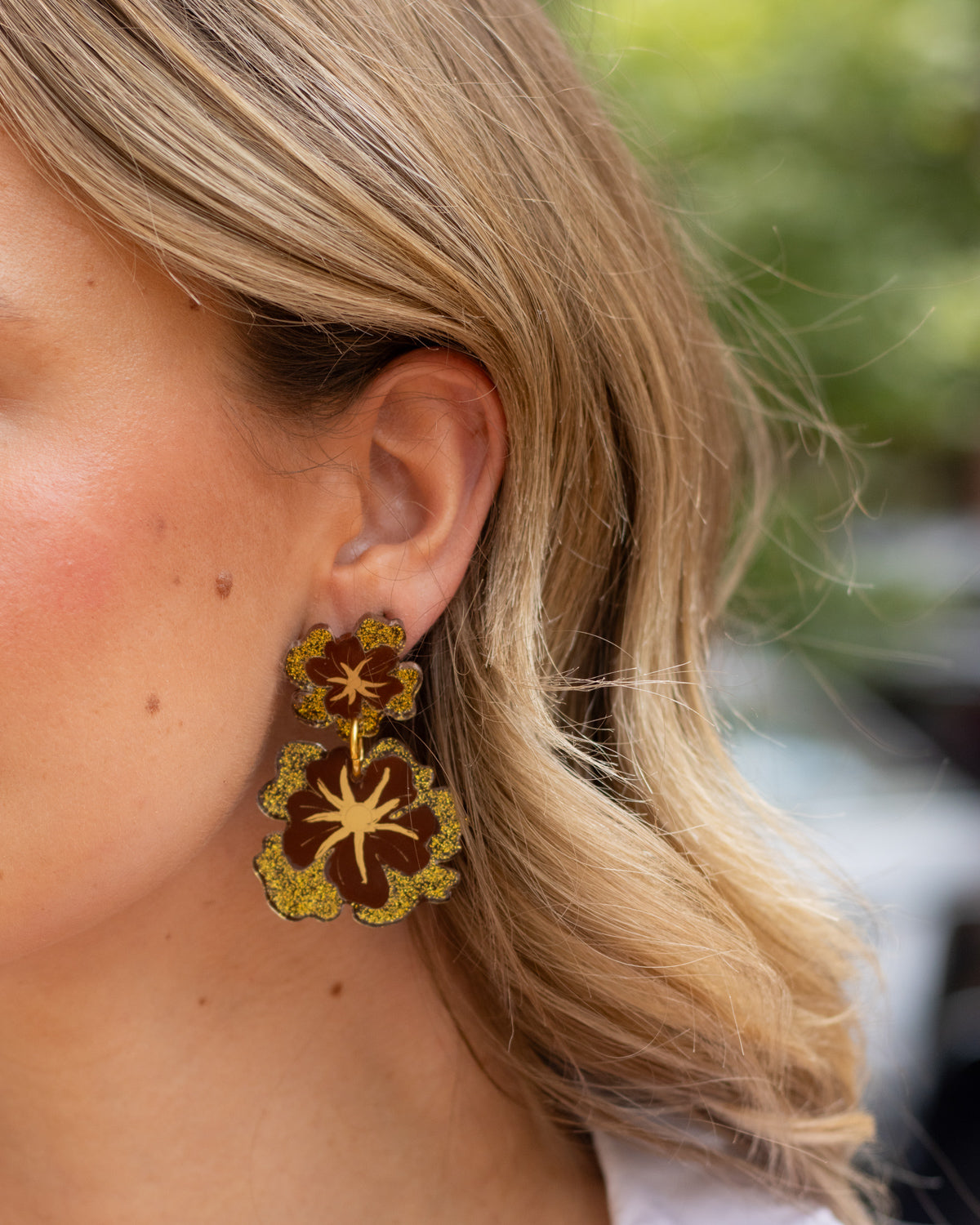 A close-up of a woman's ear wearing a pair of dangling earrings with a gold and dark brown floral design.