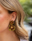A close-up of a woman's ear wearing a pair of dangling earrings with a gold and dark brown floral design.