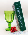 Tinseltown puzzle box standing beside a green glass goblet and red berries