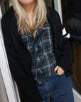 Close up of woman wearing the Pink Pineapple Après Ski black varsity cardigan over a ruffled flannel Dôen top and jeans standing in a doorway.