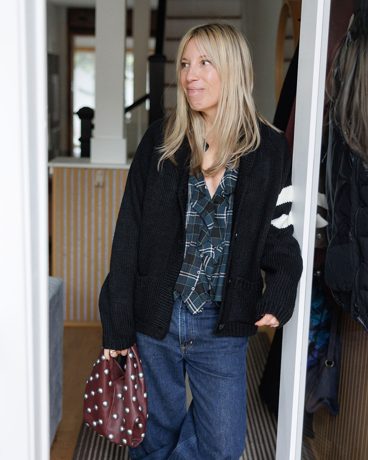 Woman wearing the Pink Pineapple Après Ski black varsity cardigan over a ruffled flannel Dôen top and jeans standing in a doorway.