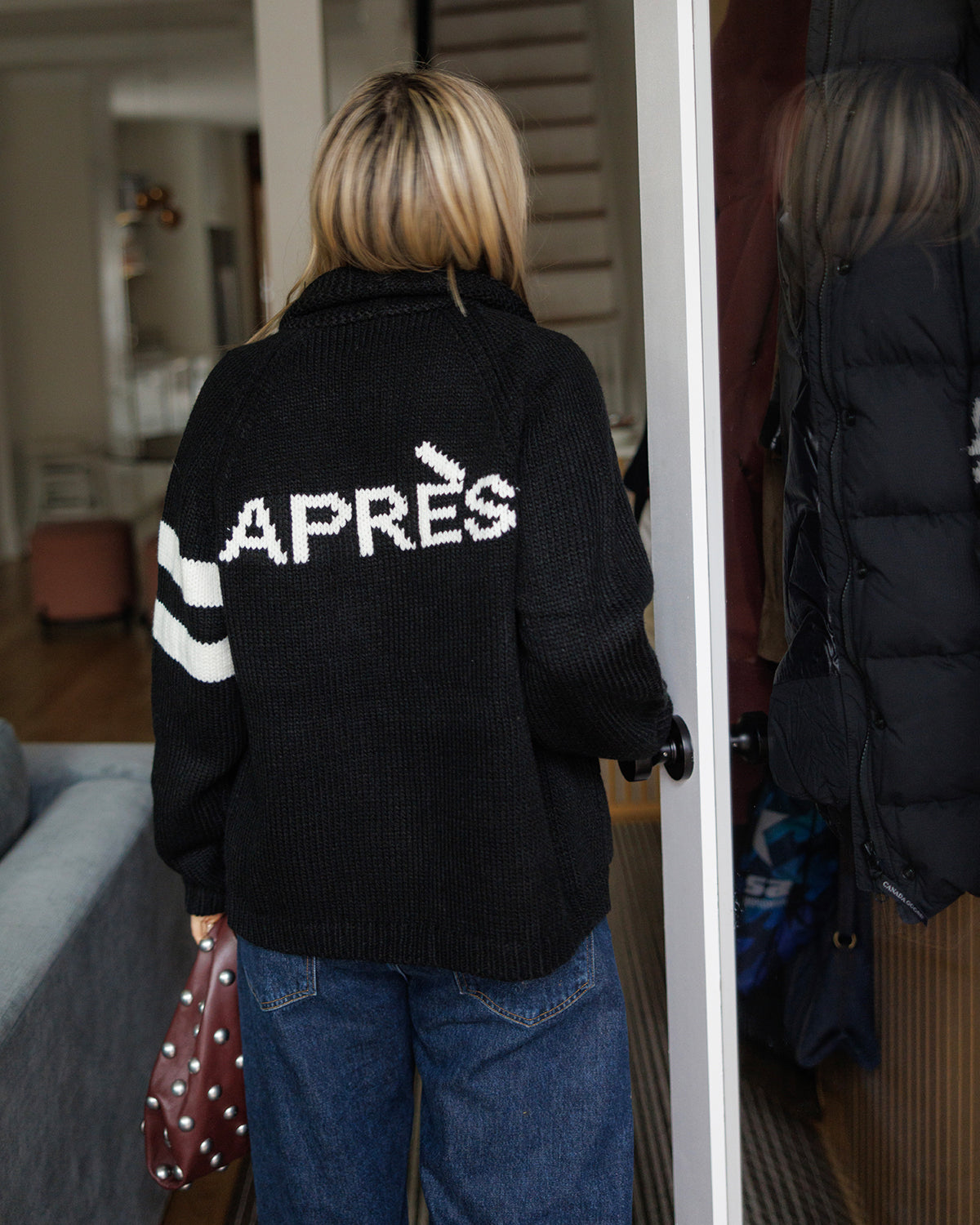 Back of a woman wearing Pink Pineapple Après ski black varsity cardigan with white stripes on one sleeve and Après graphic text on the back.