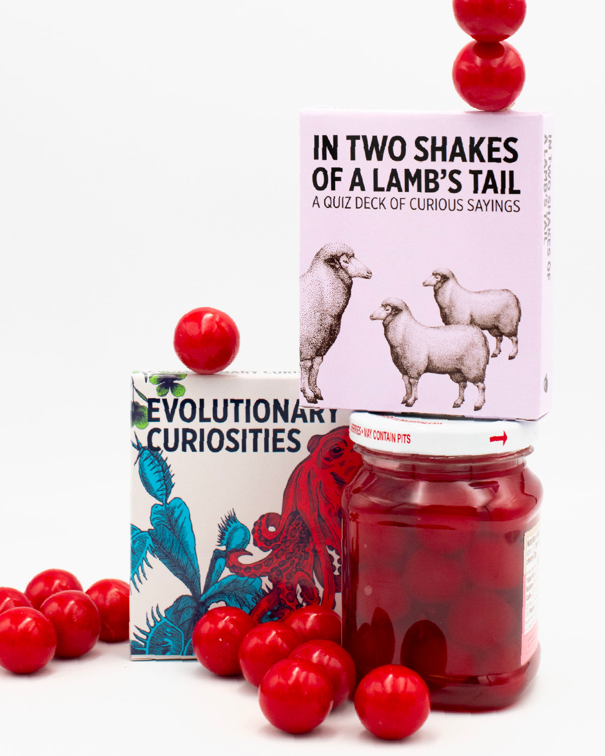 Photo of the In Two Shakes of a Lamb’s Tail sayings deck stacked on a jar of red maraschino cherries, next to the Evolutionary Curiosities knowledge card deck with festive holly berry decorations