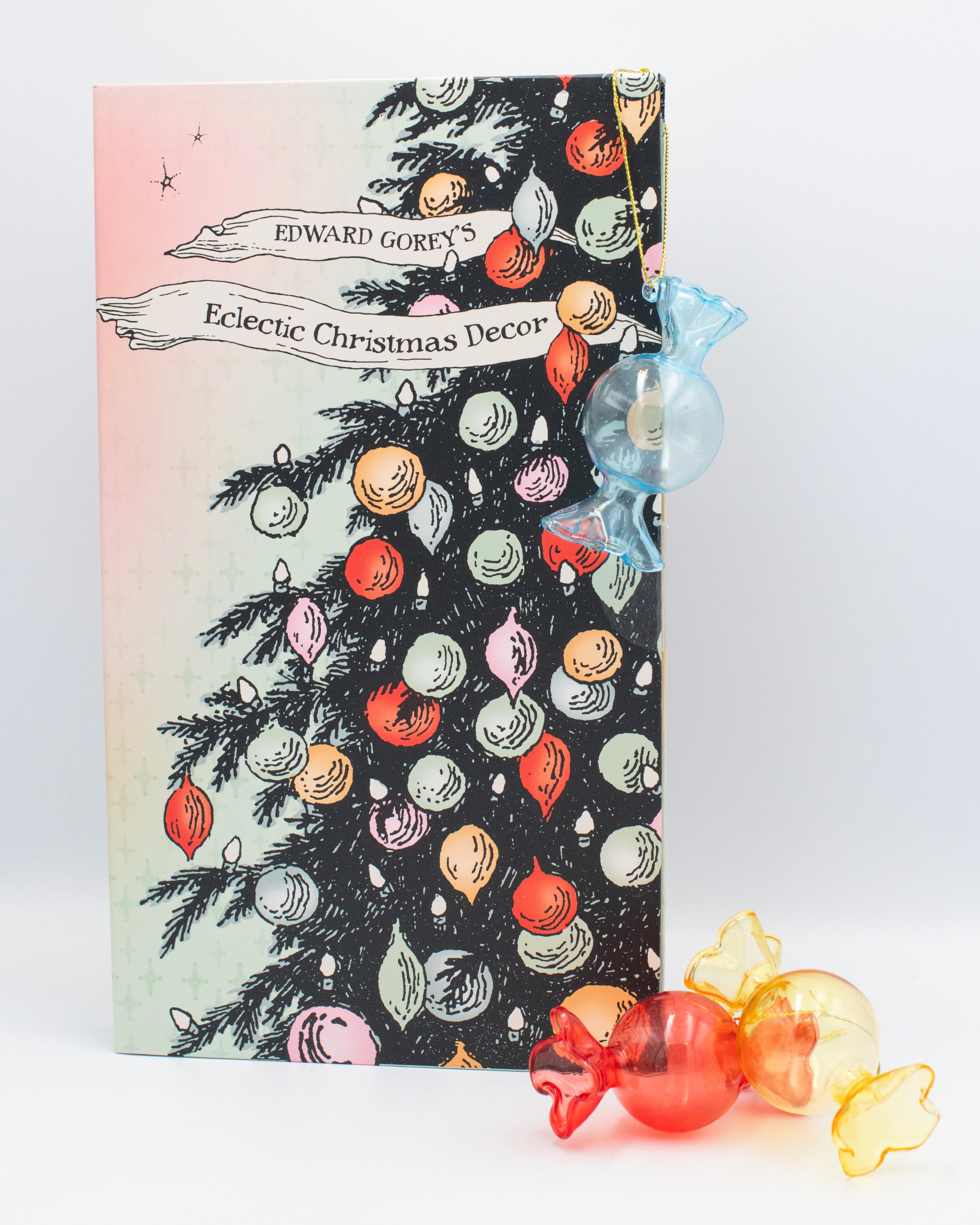 Edward Gorey's Eclectic Christmas Decor craft box feautring an illustration of a Christmas tree, ornaments and holiday lights beside glass blown candy ornaments.