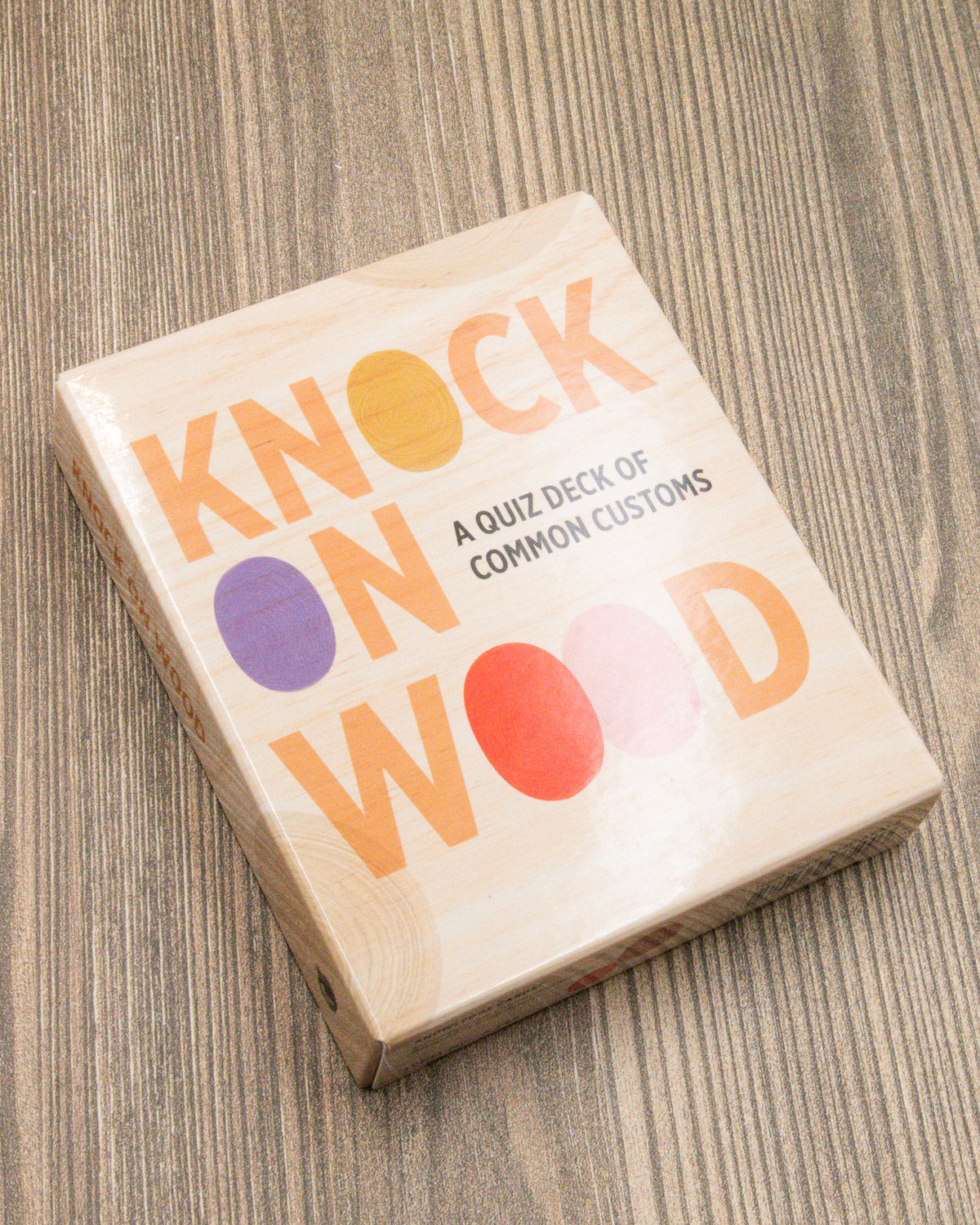 Knock on Wood card deck box with peach and purple lettering on a wood table