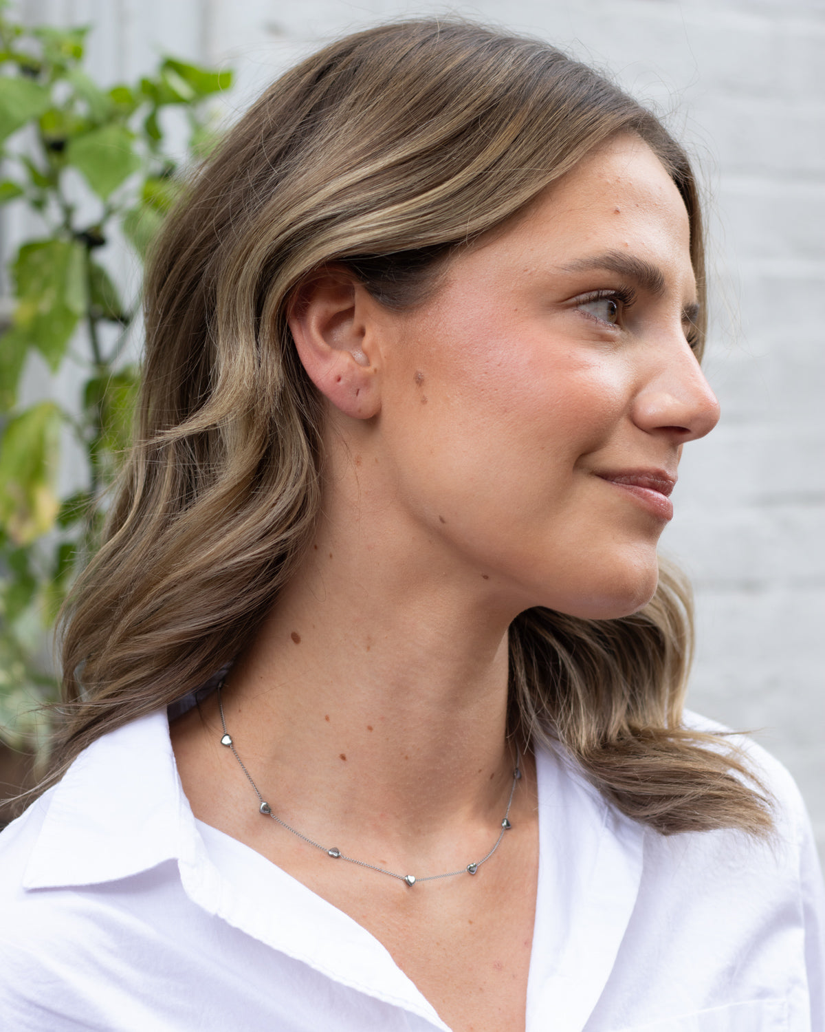 A woman in a white collared shirt wearing a delicate silver chain necklace with small heart-shaped charms, shown in a profile view against a light gray brick wall background.