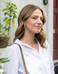 A woman with shoulder-length wavy brown hair wearing a white button-down shirt and a delicate silver necklace, carrying a brown shoulder strap, set against a soft-focus background of a white brick wall and a red door.