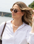 A woman outdoors wearing oval dark sunglasses, a white button-up shirt, and a pearl necklace, with a bridge and water blurred in the background.