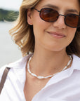 A close-up of a woman wearing dark brown tinted sunglasses with a black frame, a white collared shirt, and a pearl shell necklace.