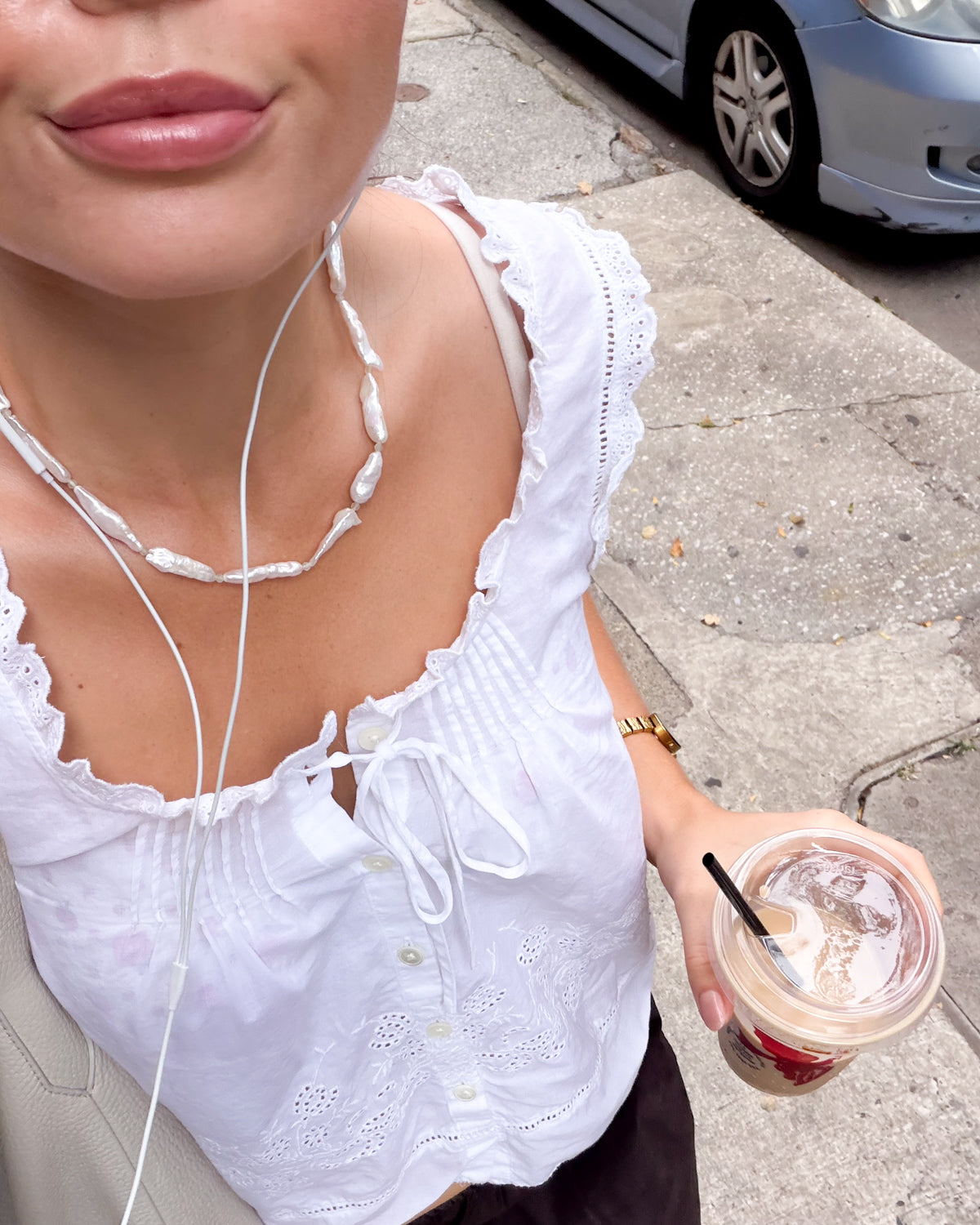 A close-up lifestyle shot of a person wearing a white eyelet lace top with ruffled trim and a front tie closure, holding an iced coffee on a city sidewalk. The person is also wearing a chunky white pearl  beaded necklace and a gold watch.