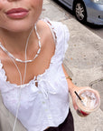A close-up lifestyle shot of a person wearing a white eyelet lace top with ruffled trim and a front tie closure, holding an iced coffee on a city sidewalk. The person is also wearing a chunky white pearl  beaded necklace and a gold watch.