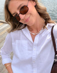 A woman wearing a white long-sleeve button-up shirt, brown sunglasses, and a beaded pearl necklace, standing on a wooden dock with water in the background.