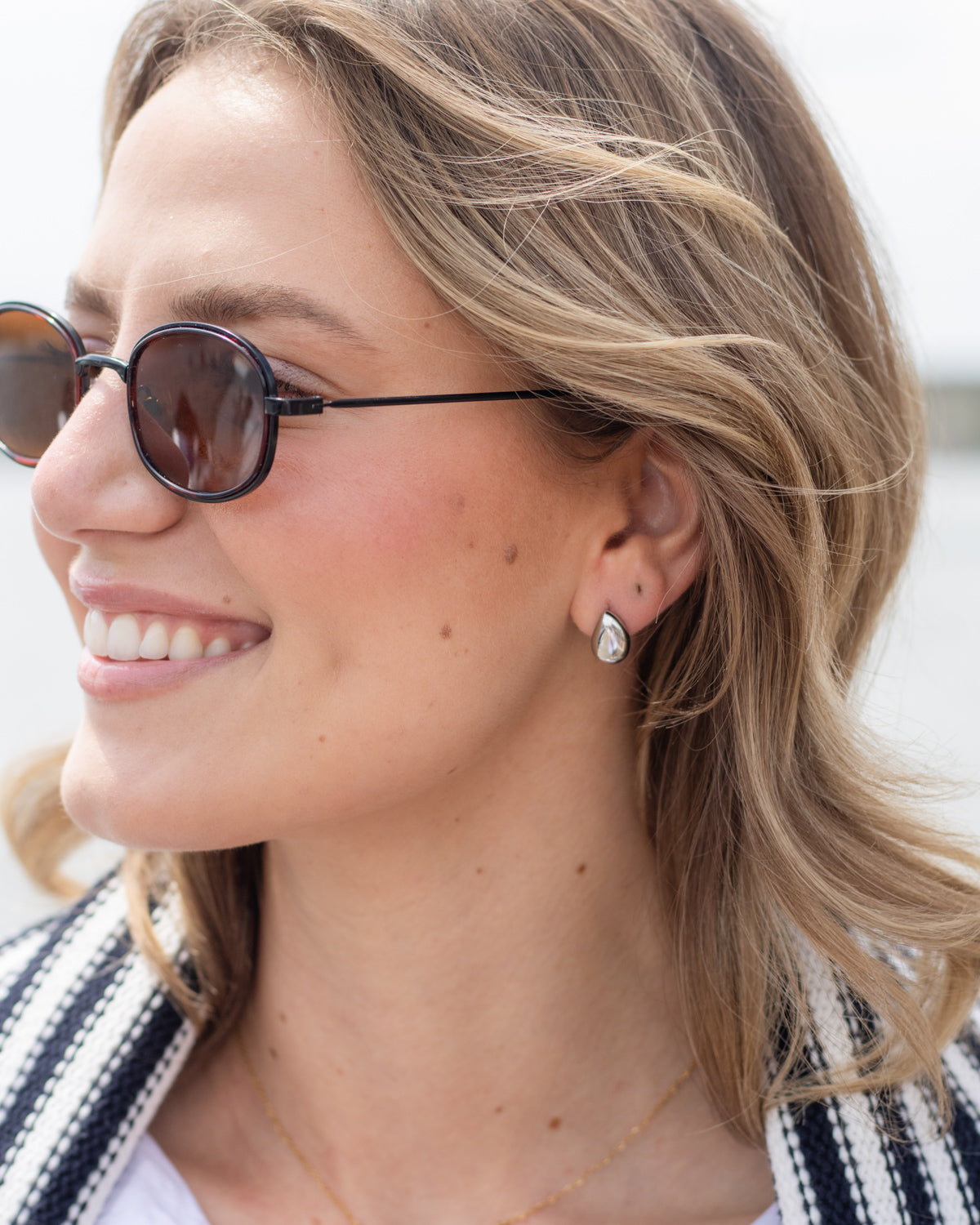 A close-up side profile of a smiling woman with blonde hair wearing round dark sunglasses and a small silver teardrop earring.