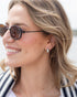 A close-up side profile of a smiling woman with blonde hair wearing round dark sunglasses and a small silver teardrop earring.