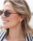 A close-up side profile of a smiling woman with blonde hair wearing round dark sunglasses and a small silver teardrop earring.