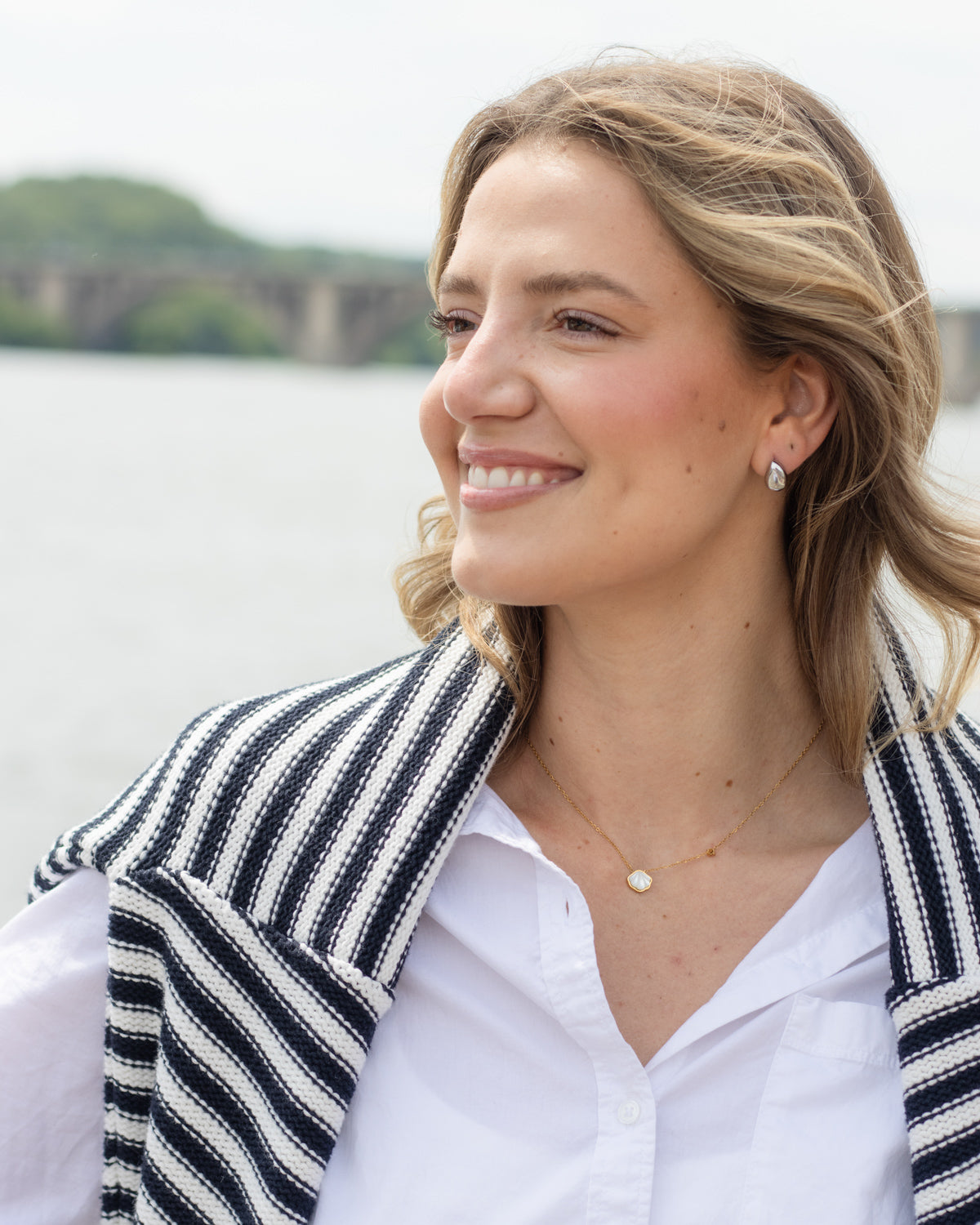 A smiling blonde woman in a white button-down shirt and a black and white striped knit cardigan, wearing a delicate gold necklace and earring, photographed outdoors with a blurred body of water and green hills in the background.
