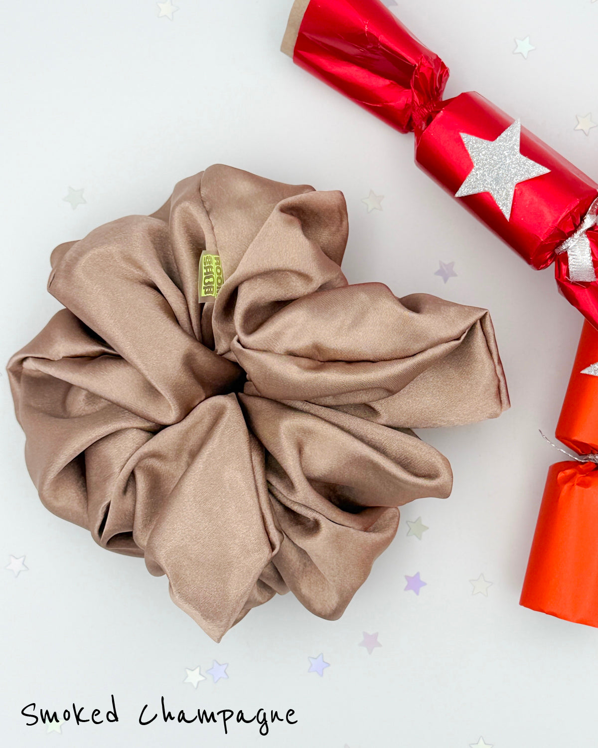 Giant, oversized satin scrunchie in smoked champagne color next to a festive red, Christmas cracker with a silver star.