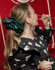 Profile of a woman with an oversized satin midnight green scrunchie over a ponytail against a red background drinking a glass of champagne.