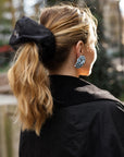Woman with blonde hair wearing an oversized black, organza scrunchie over a ponytail, wearing a black jacket.