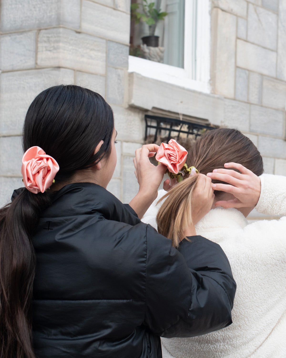 Two young women in pontyails adjusting pink satin rosette scrunchies in front of a stone building.