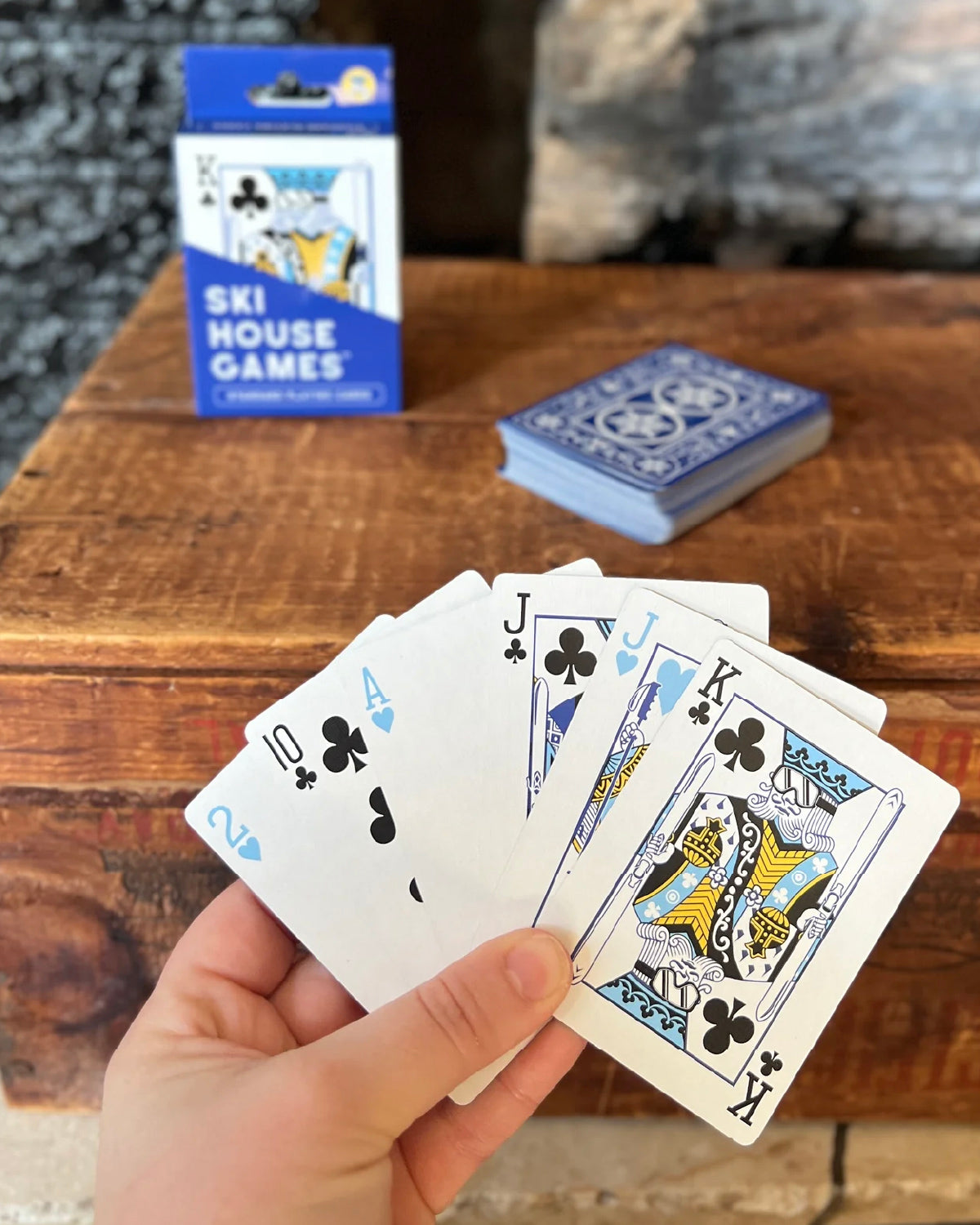 Hand holding a fan of Ski House Games cards with the blue box and deck on a wood table